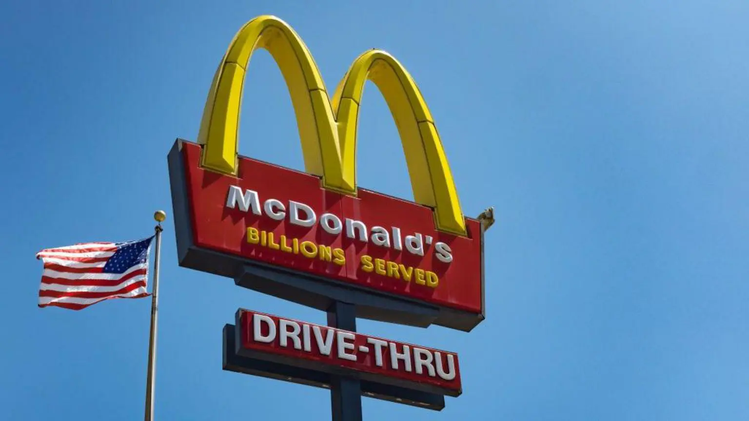 Знак McDonald's «Золоті арки» на тлі блакитного неба з прапором США, що майорить унизу ліворуч