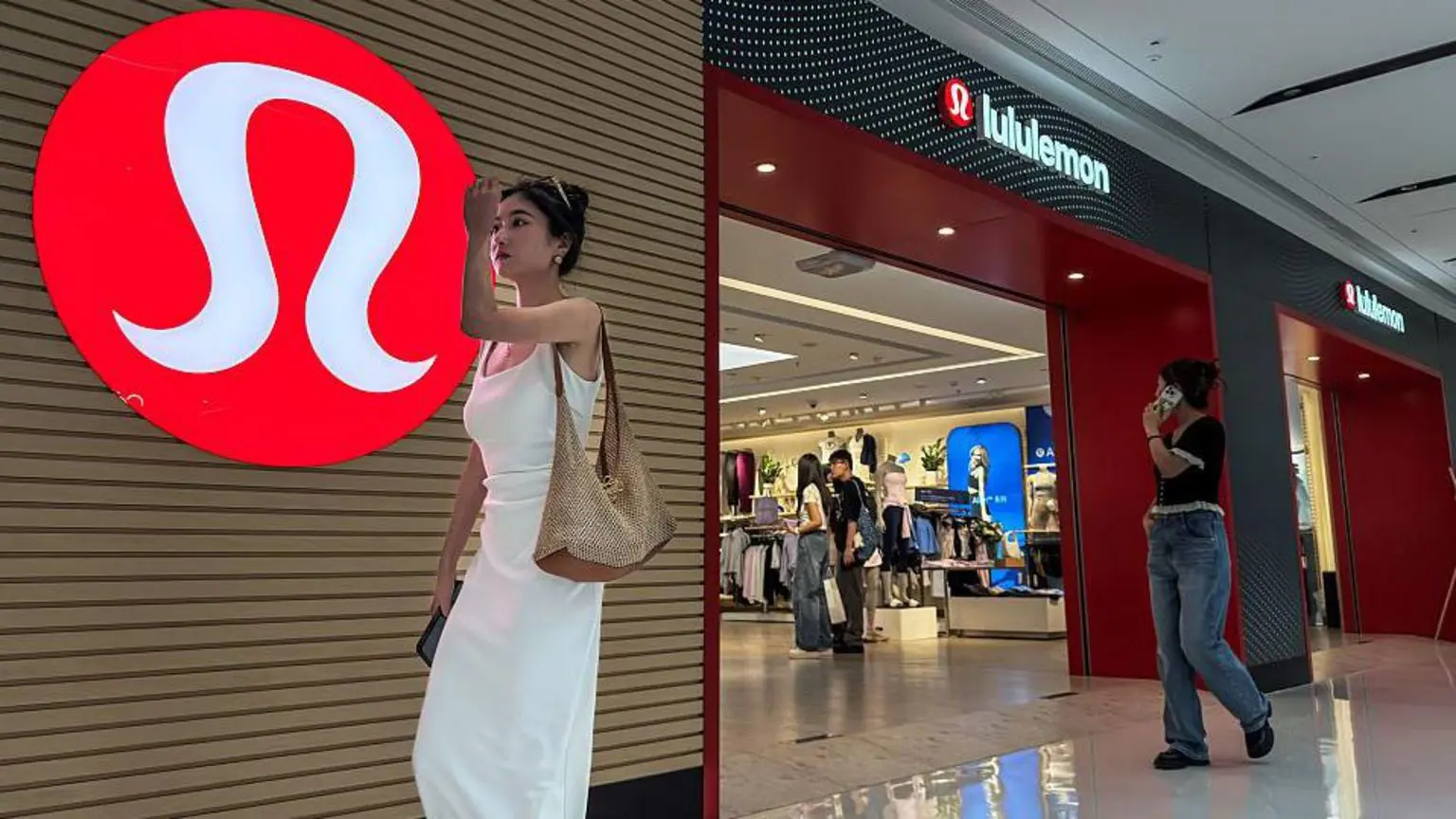 Жінка в білій сукні проходить повз магазин Lululemon із фірмовим червоним логотипом, який видніється на стіні, поки покупці переглядають магазин, 4 червня 2025 року в Чунціні, Китай.