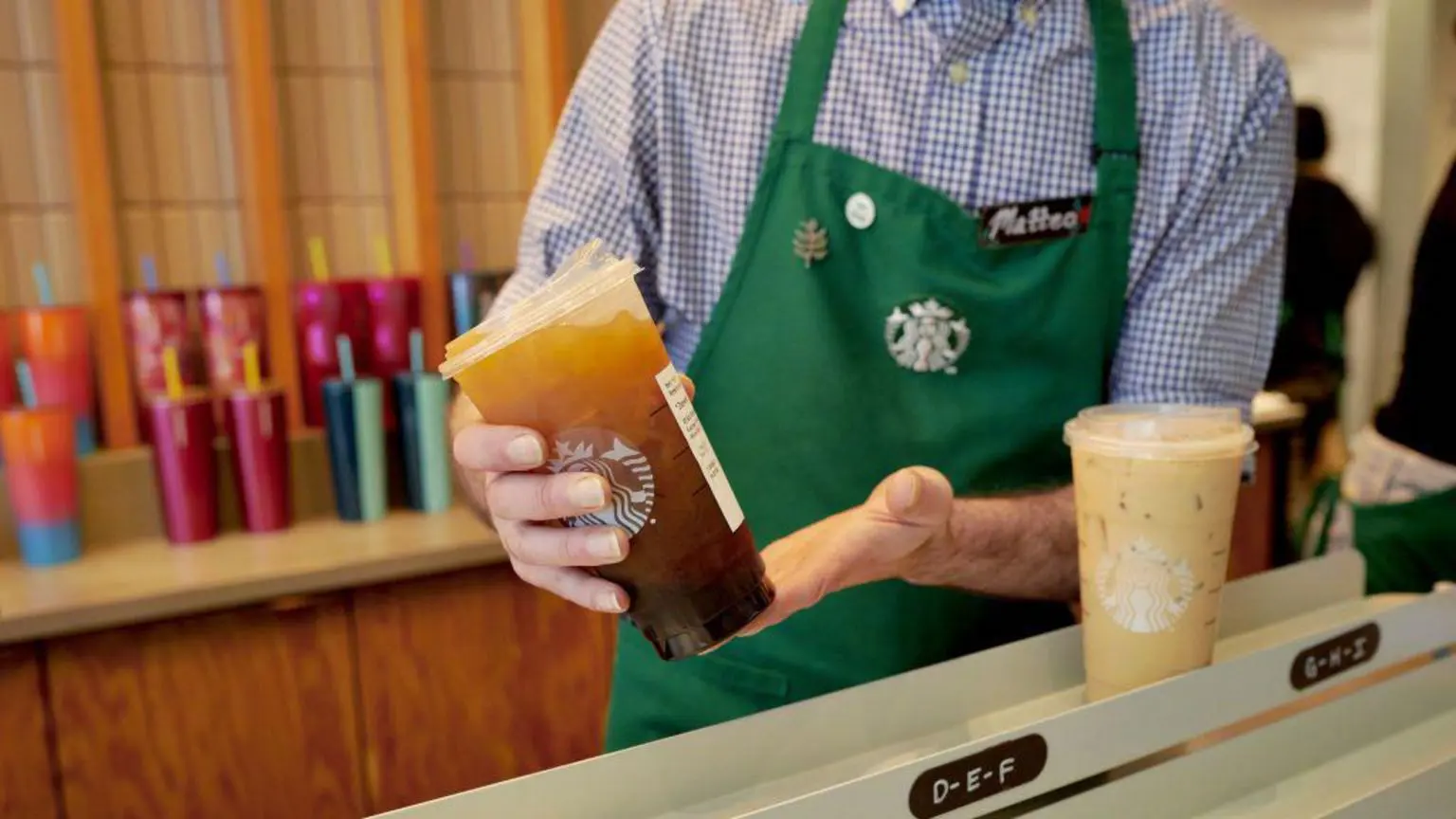 Бариста розміщує каву з льодом у зоні мобільного прийому в Starbucks у Нью-Йорку, США. Бариста одягнений у синьо-білу картату сорочку з зеленим фартухом із логотипом Starbucks у центрі.