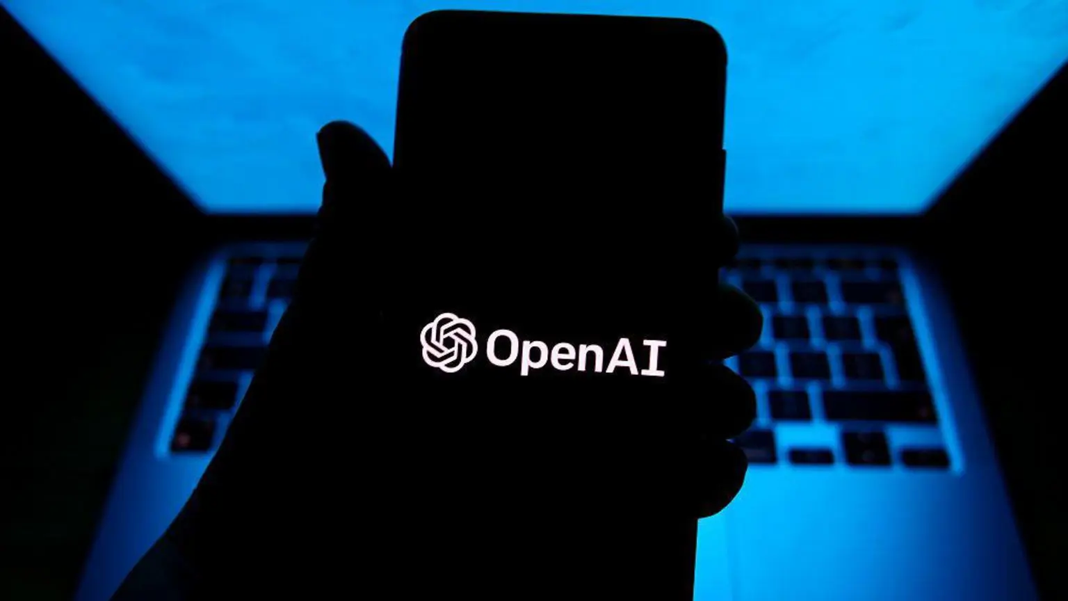 Людина тримає телефон із програмним забезпеченням OpenAI у темній кімнаті з комп’ютером за рукою