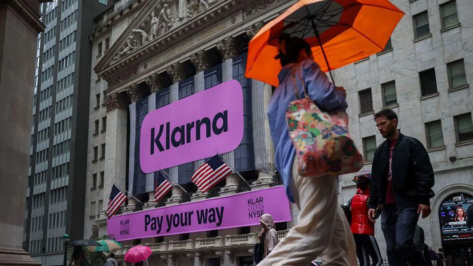 Знак кольору фусії з написом Klarna та Pay your way прикриває Нью-Йоркську фондову біржу, поки люди з парасольками проходять повз неї в день дебюту на Нью-Йоркській фондовій біржі (NYSE) у Нью-Йорку, США, у середу, 10 вересня 2025 року.