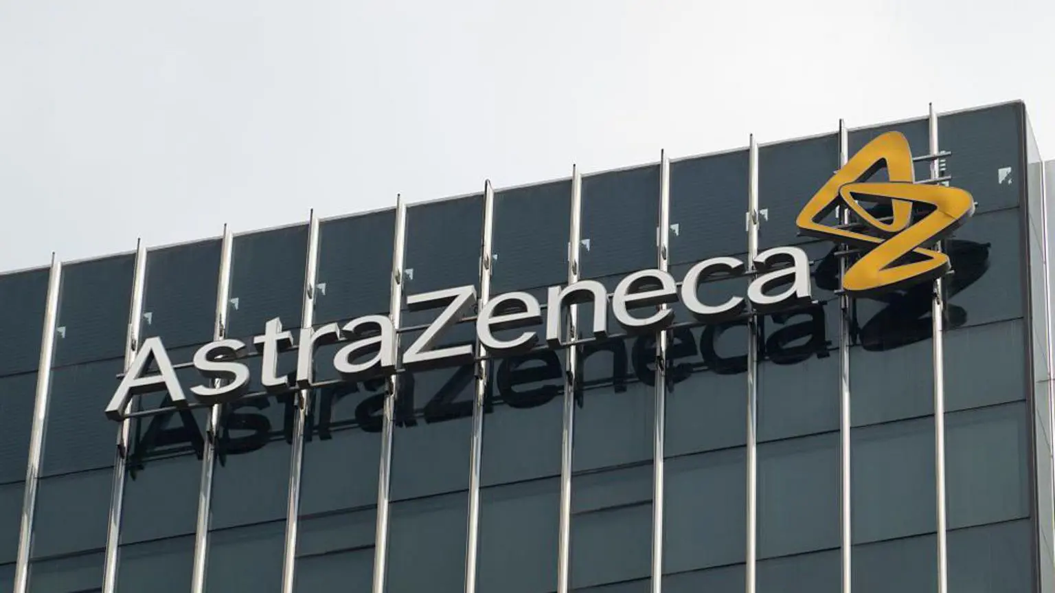 Логотип фармацевтичної компанії Astrazeneca на боці офісної будівлі з темними непрозорими вікнами. За будівлею сіре небо.