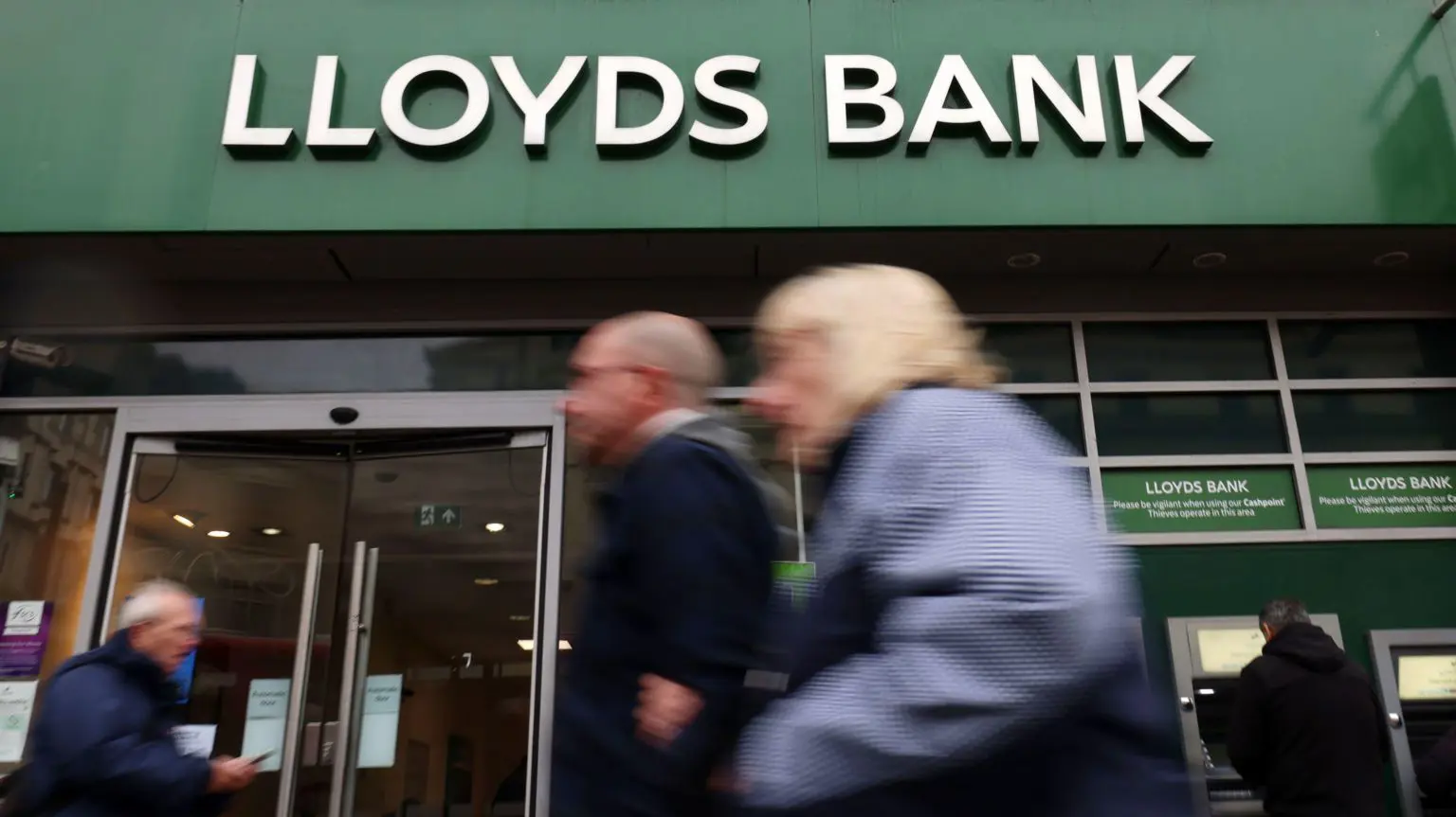 Знімок фасаду банку Lloyds – його фірмова назва написана білими літерами над банком на характерному зеленому фоні – на передньому плані зображено кілька людей, що йдуть, і вони розфокусовані, що привертає увагу до назви банку – також можна побачити людину, яка знімає гроші з банкомата