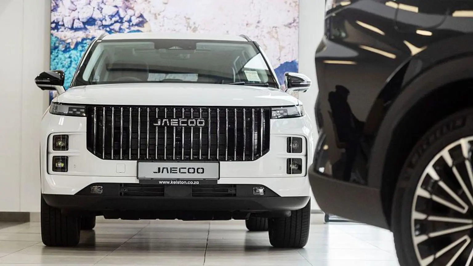 Білий позашляховик стоїть на підлозі автосалону, на передньому плані — задня частина чорного позашляховика.