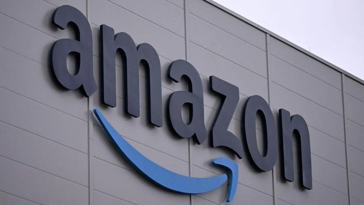Логотип Amazon, який являє собою світове слово «Amazon» з його фірмовою стилізованою стрілкою під ним, на стіні сірої будівлі з легким похмурим небом на задньому плані