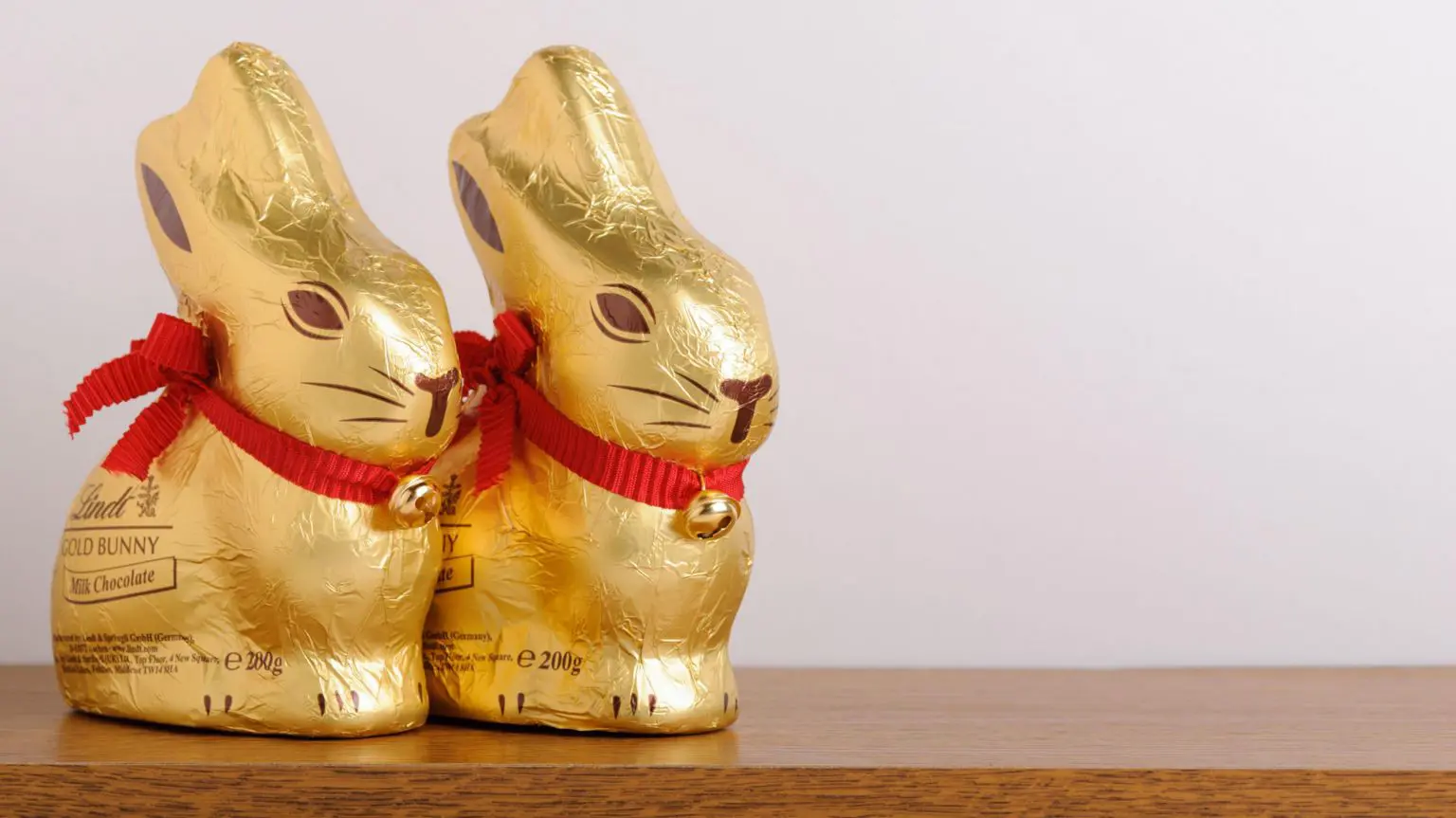 Два великодні шоколадні кролики Lindt поруч один з одним на дерев'яному столі з порожньою бежевою стіною позаду. Кролики загорнуті в золоту фольгу та мають червону стрічку на шиї з дзвіночком.