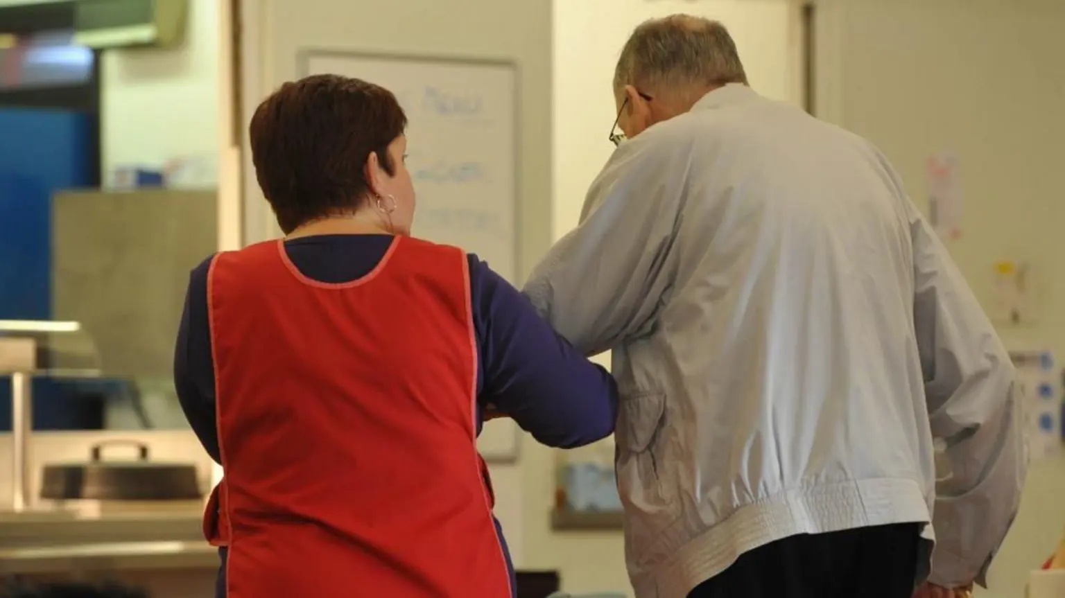 Жінка-доглядальниця в червоному фартусі допомагає літньому чоловікові в сірій куртці