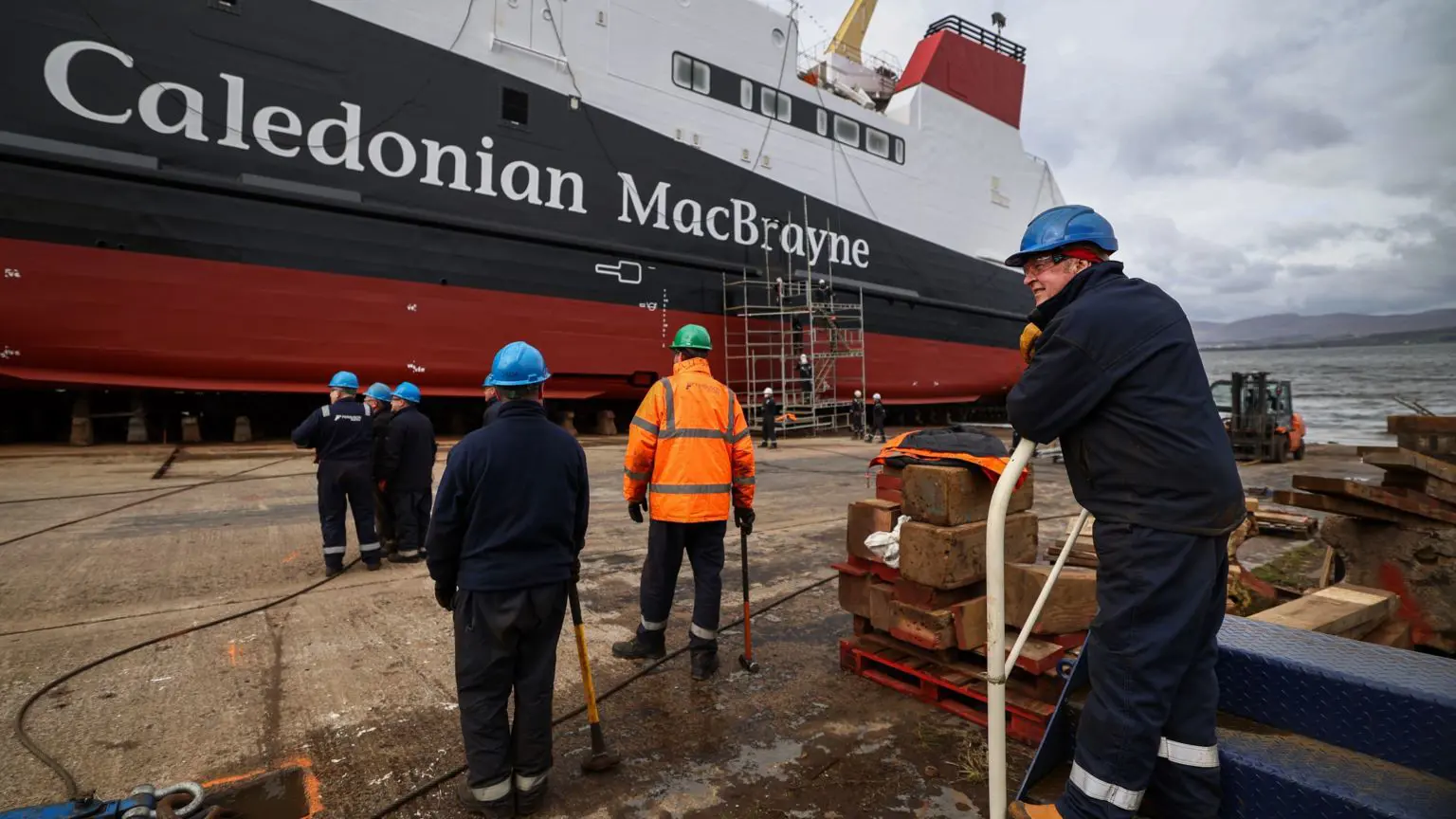 Робітники проводять останні приготування перед спуском на воду судна MV Glen Rosa у квітні 2024 року. На передньому плані спиною до камери видно шістьох робітників. Вдалині видно біло-чорно-червоний пором з написом Caledonian MacBrayne на боці.
