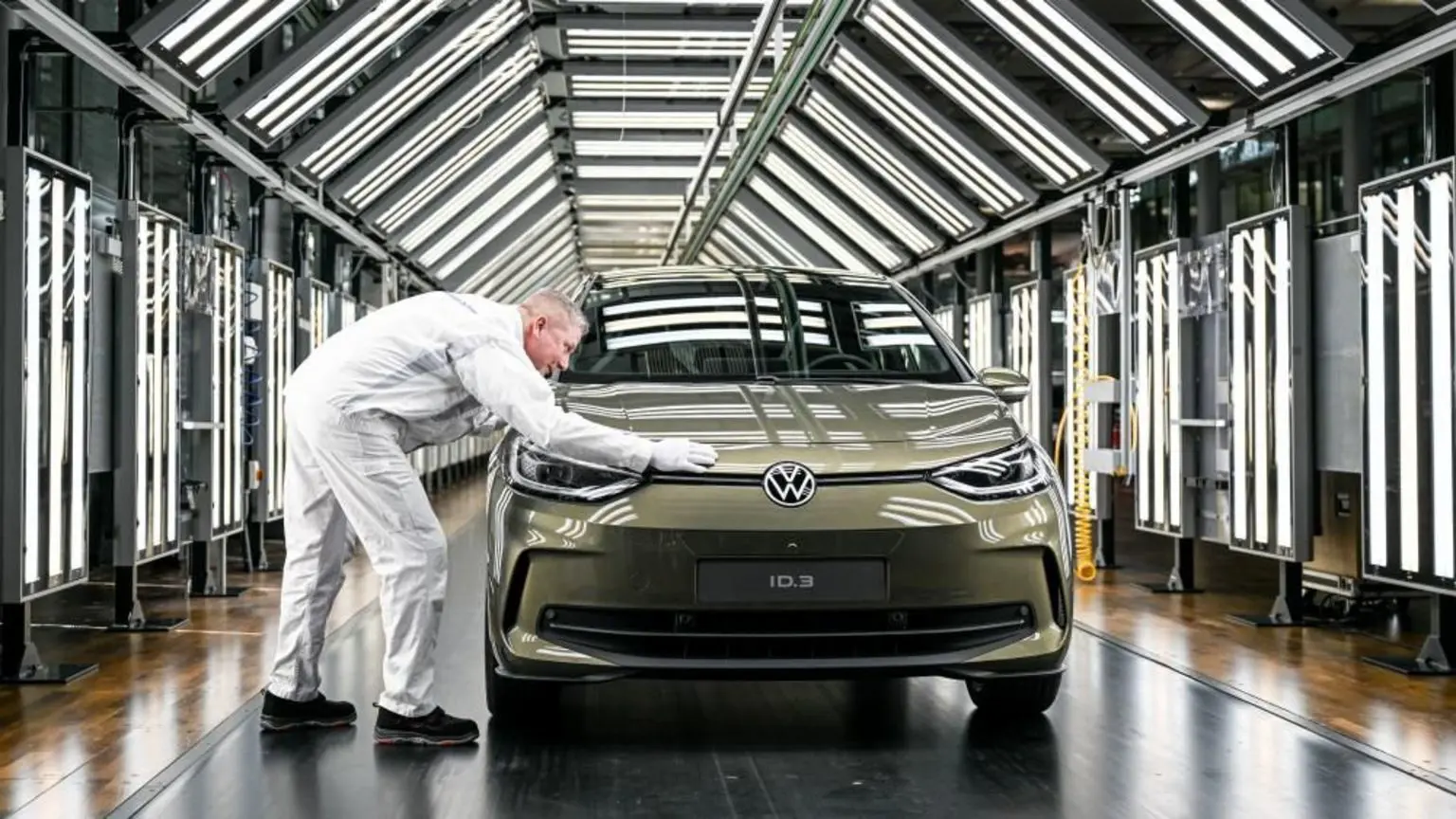 Співробітник Volkswagen демонструє автомобіль Volkswagen ID.3 під час прес-презентації. Він тримає руку над автомобілем моховисто-зеленого кольору, припаркованим у тунелі, заповненому білими світлодіодними панелями.